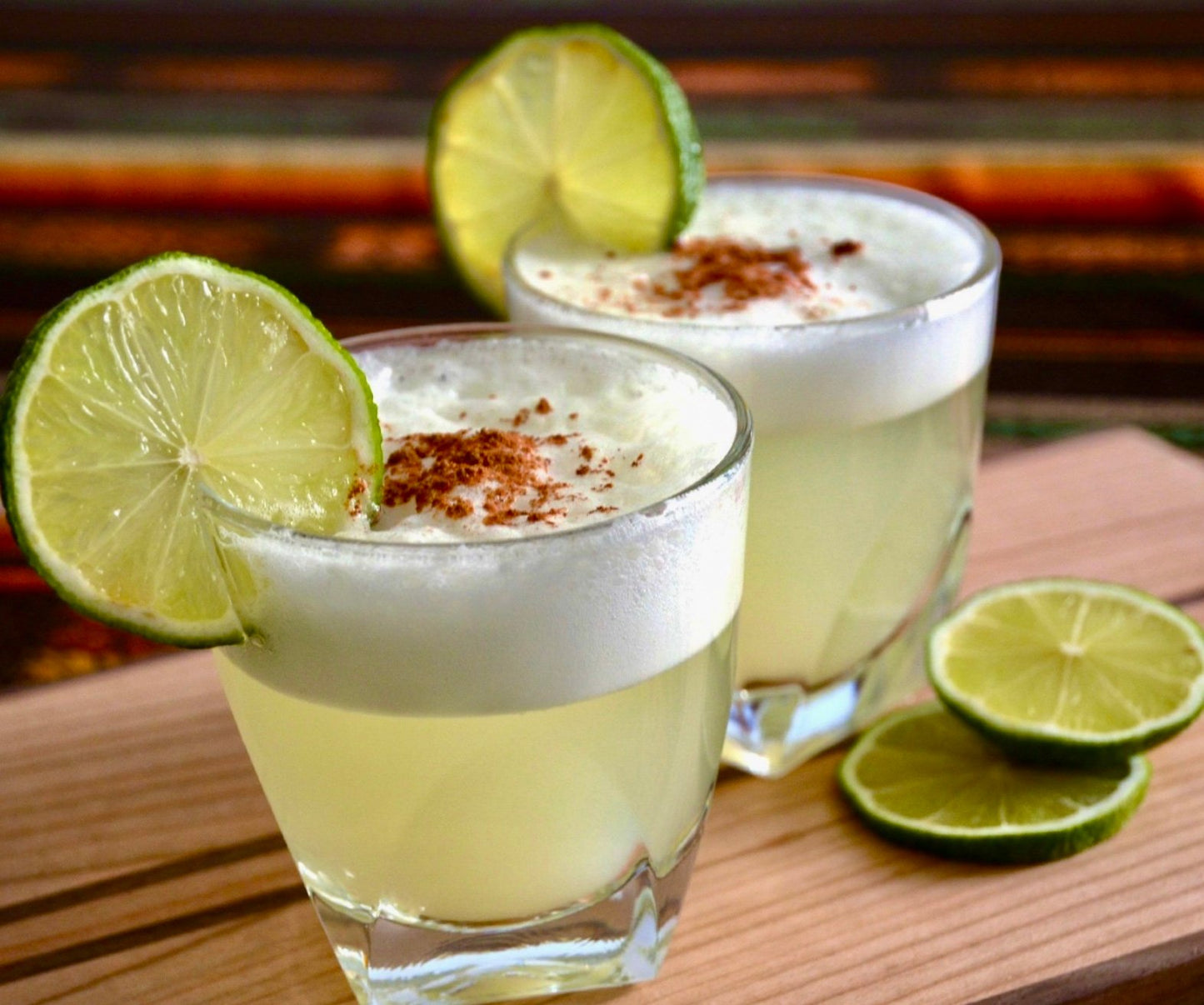 Two pisco sour cocktails garnished with lime slices on a wooden board, showcasing their frothy texture.