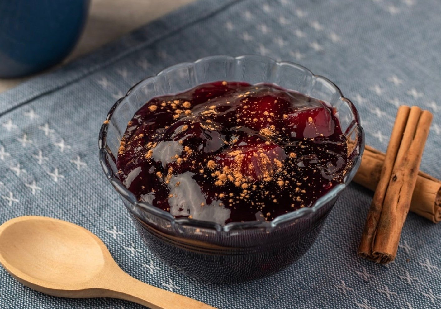 Bowl of Mazamorra Morada Umsha dessert topped with fruit and spices, on a blue textured tablecloth.